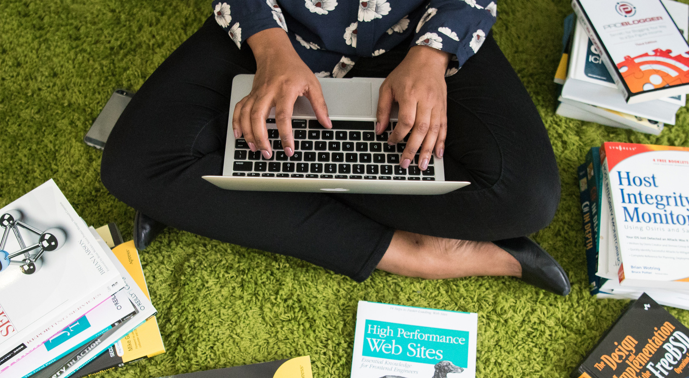 A woman sitting on a green rug cross legged and typing with Mac laptop.