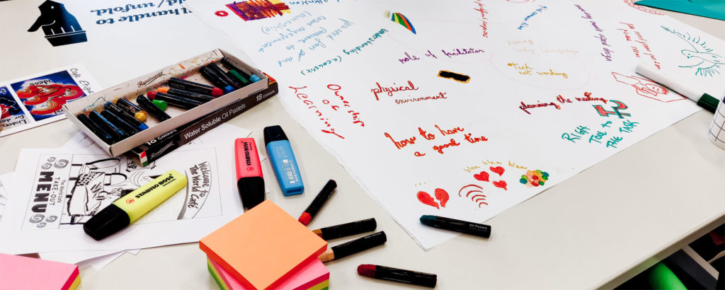 Stabilo markers, crayons and post-its spread on the table with a colorful text mindmap on the background.