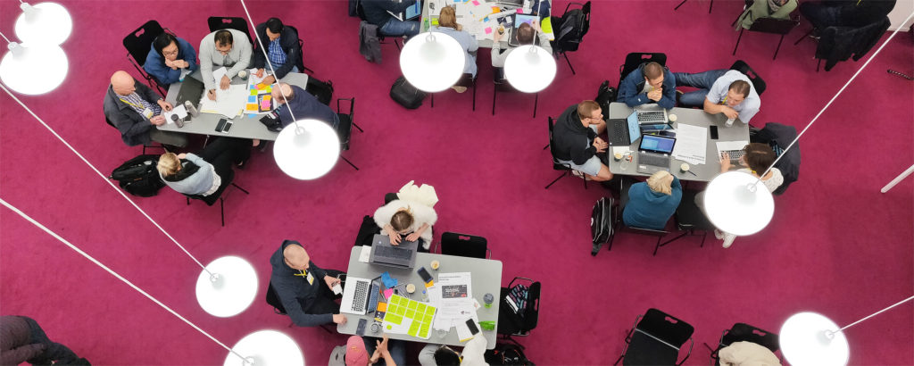 Students doing group work with laptops on the tables downstairs of the Learning Centre with fuchsia carpet and white lamps showing in the background