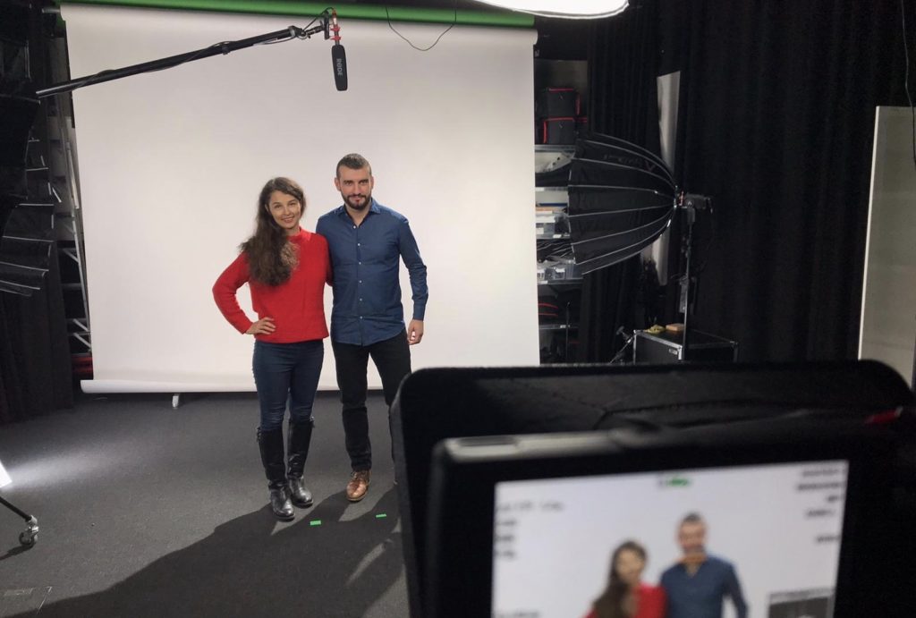 A woman and a man standing in front of a green screen and being filmed.